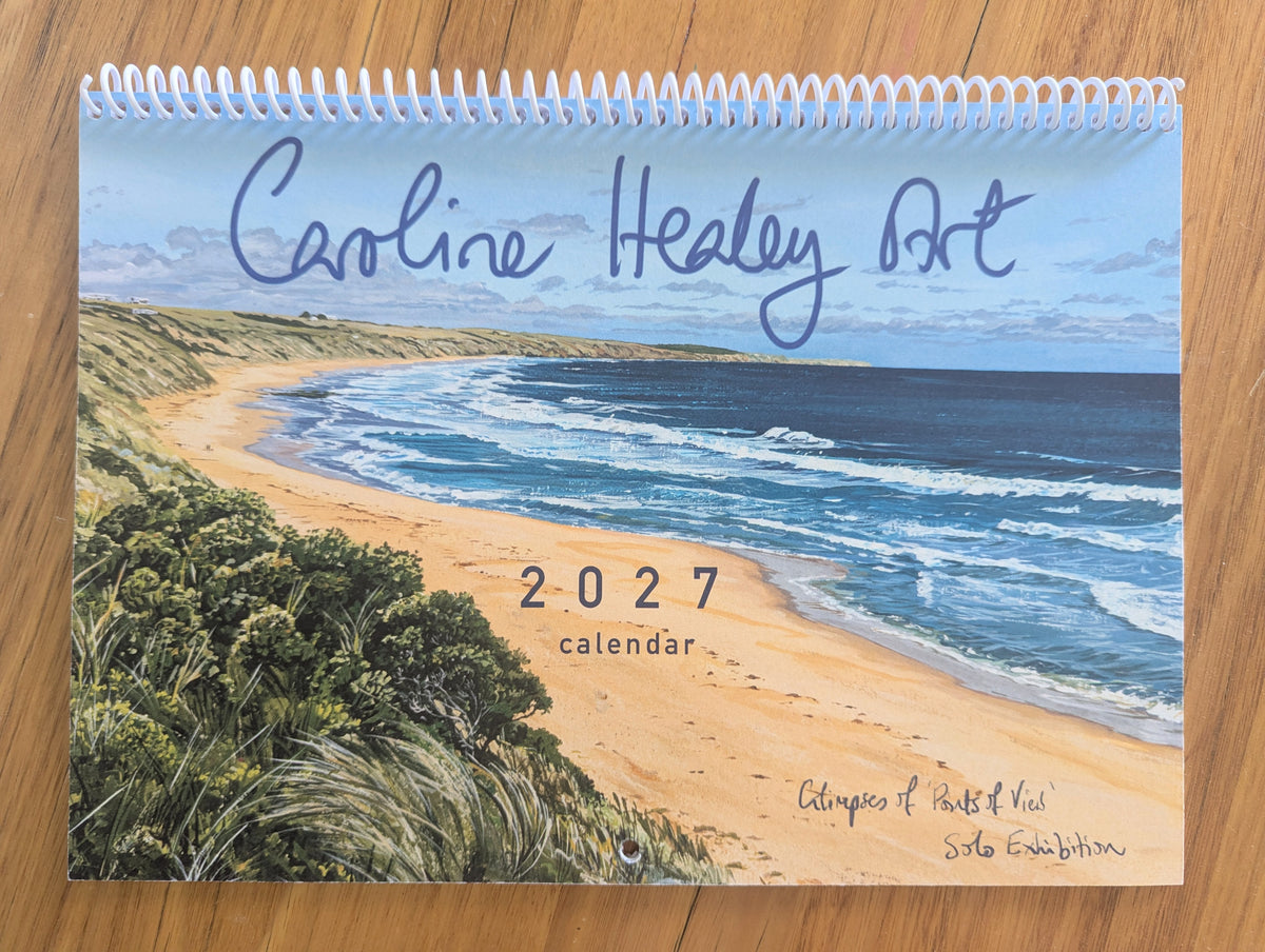 A spiral-bound 2027 calendar featuring an artistic beach scene with blue ocean waves, sandy shore, and green vegetation. The cover has the title 'Caroline Healy Art' in blue script and '2027 calendar' printed near the center, placed on a wooden surface.