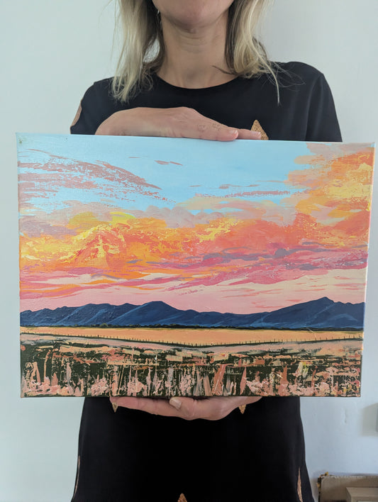 Caroline the artist holding up a canvas painting of the Grampians. A landscape at sunset, with mountains in the background and vibrant orange, pink, yellow, and blue sky.