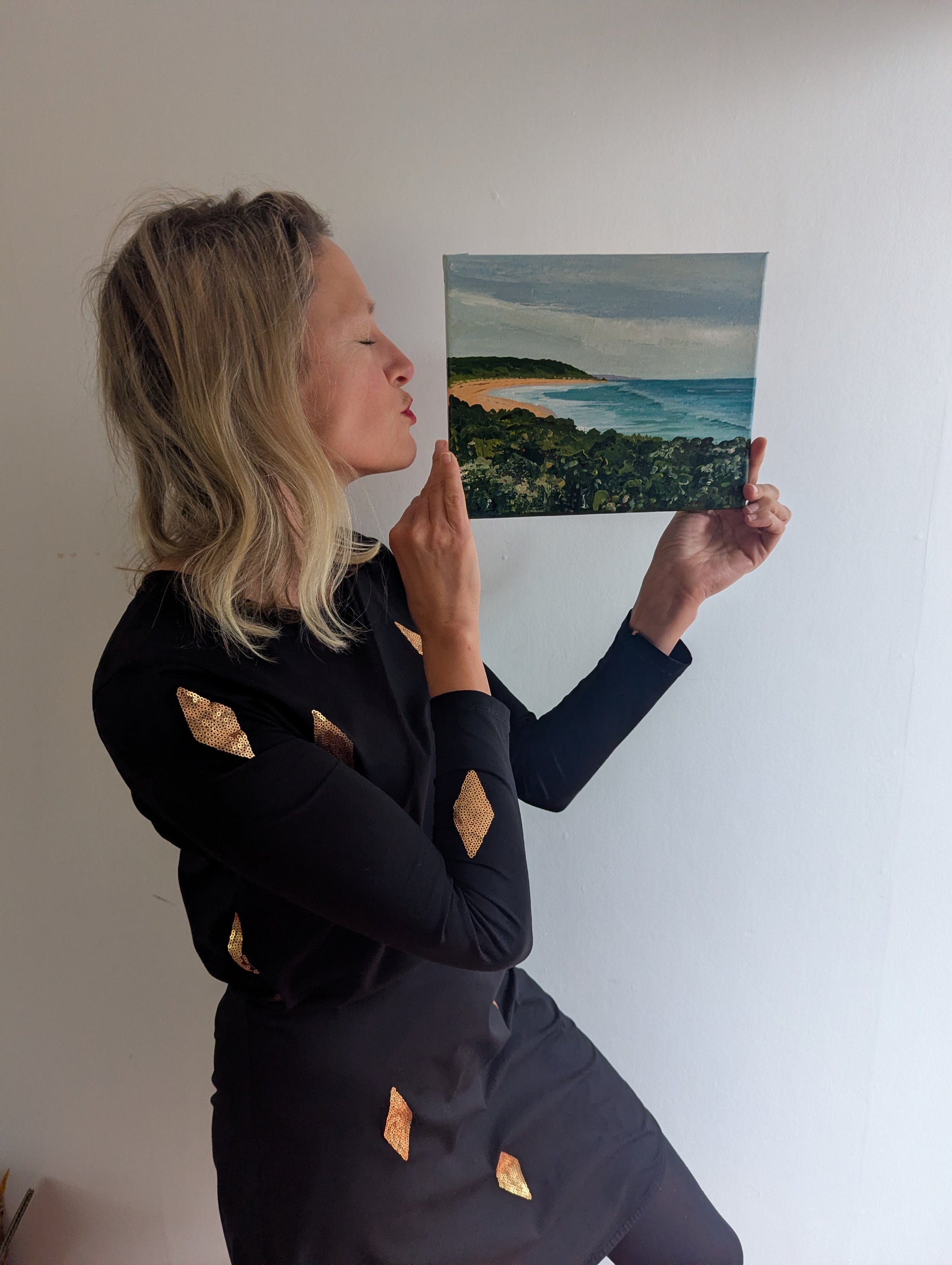 A woman in a black long-sleeve dress with gold geometric accents holds up a landscape painting of a beach and ocean against a plain white wall.